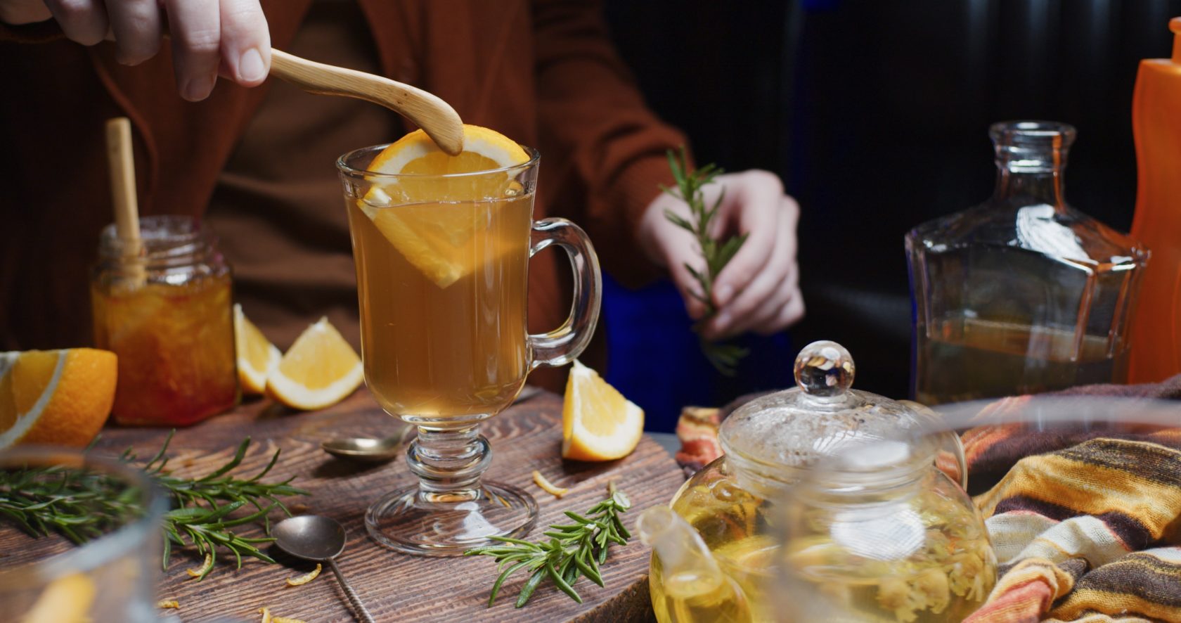 Human hands are preparing hot toddy with orange, rosemary and chamomile tea. Medium view